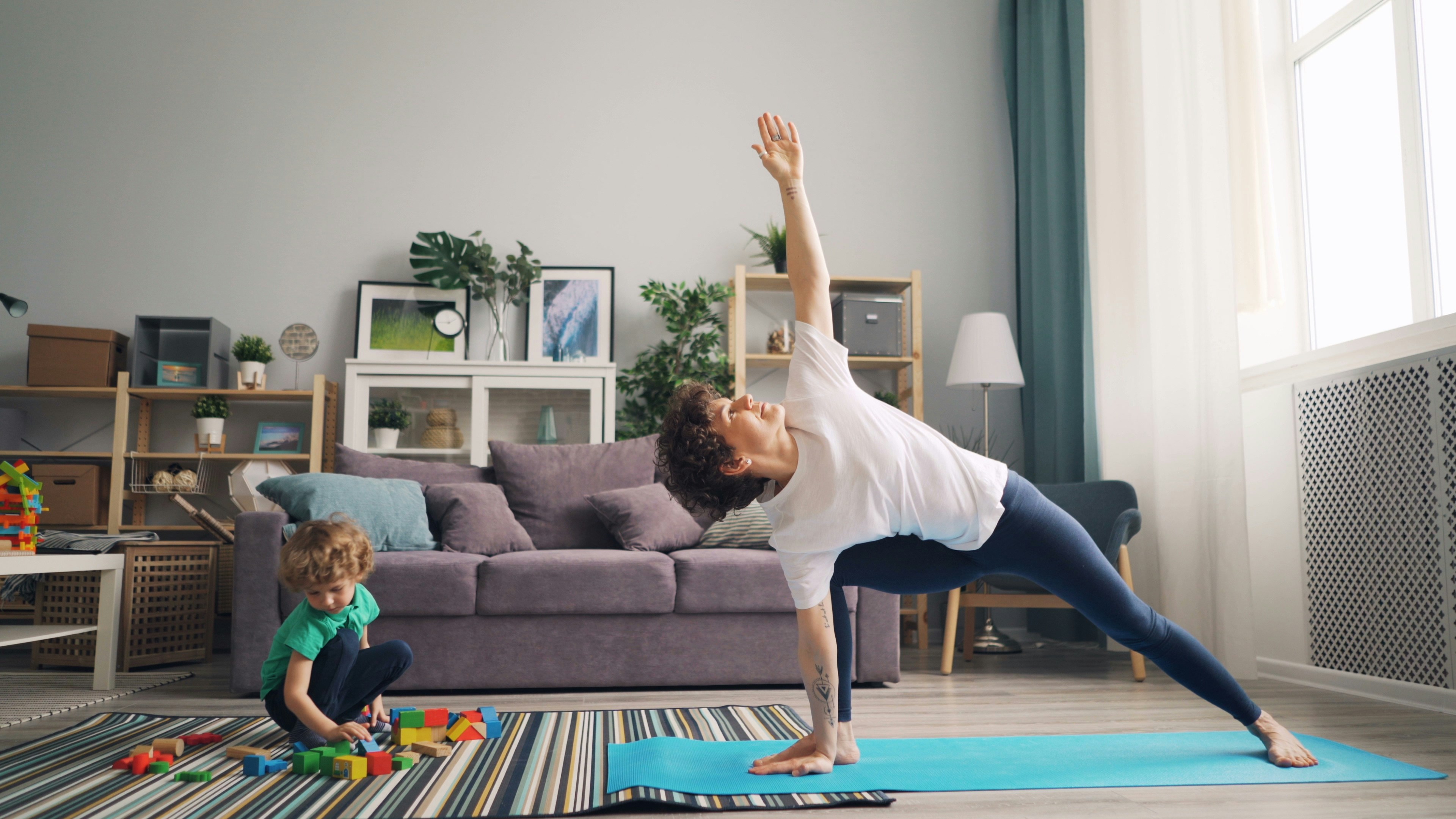 Woman practicing yoga in her living room while her young child plays with blocks nearby